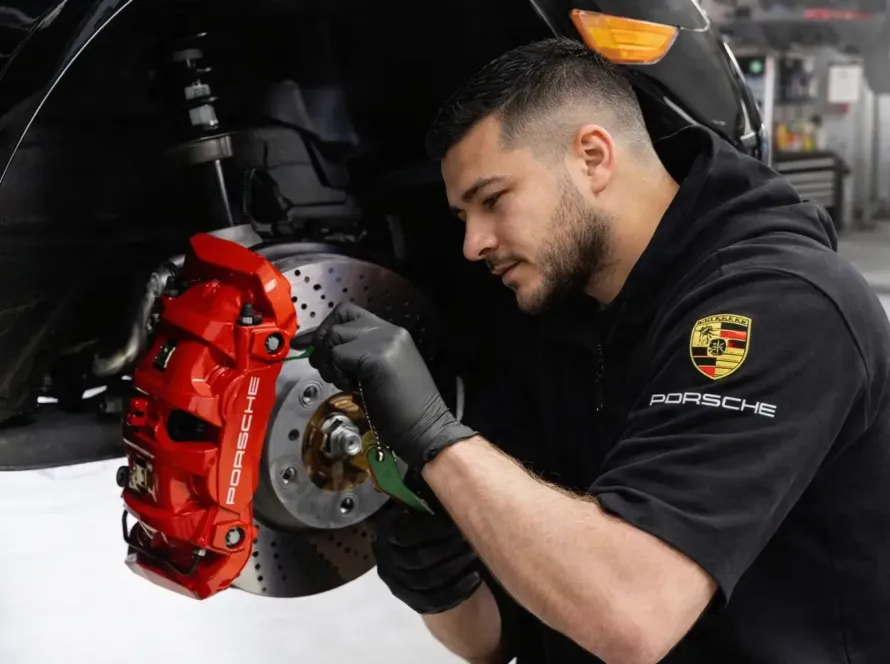 Mecânico especialista com uniforme Porsche inspecionando e revisando pinça de freio vermelha em veículo Porsche preto elevado na oficina Premium Car em Porto Alegre