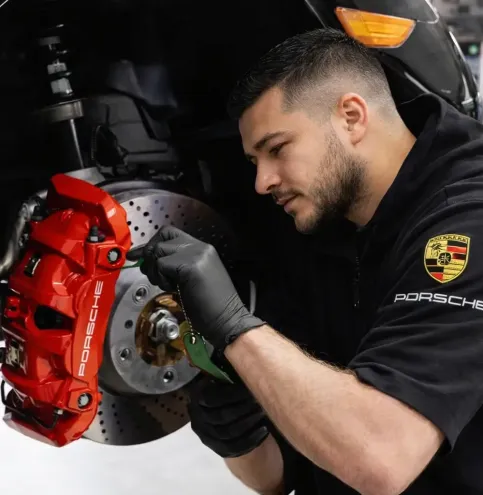 Mecânico especialista com uniforme Porsche inspecionando e revisando pinça de freio vermelha em veículo Porsche preto elevado na oficina Premium Car em Porto Alegre